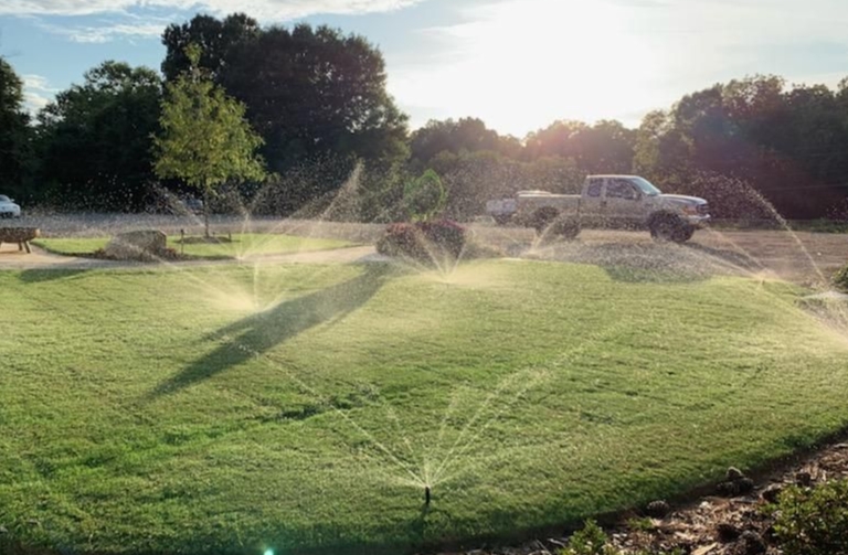 Sprinkler zones running in the evening light across a lawn, a work truck parked behind the treeline