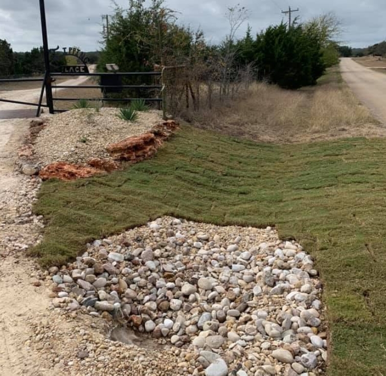 Ranch gate entrance with fresh sod laid either side and a wide river-rock drainage bed running down to the caliche road
