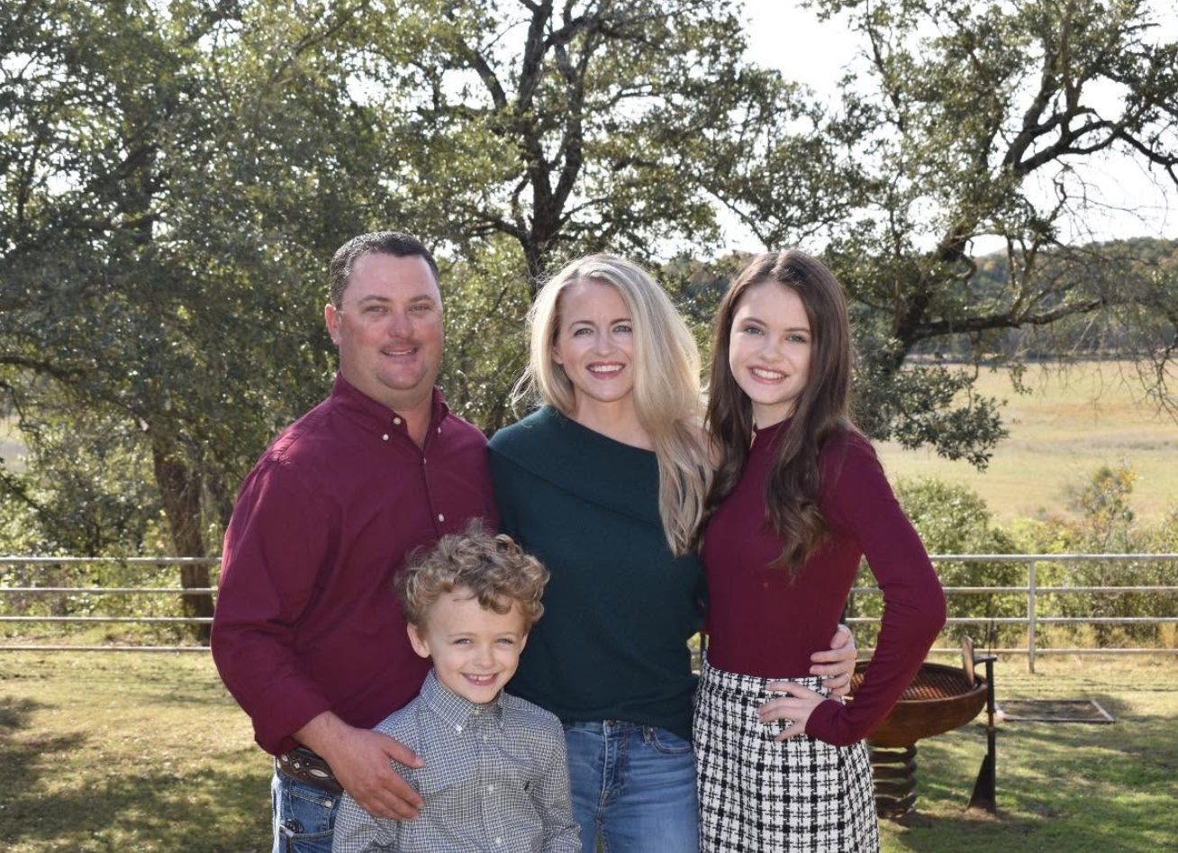 A family of four standing together under live oaks on open Texas pasture